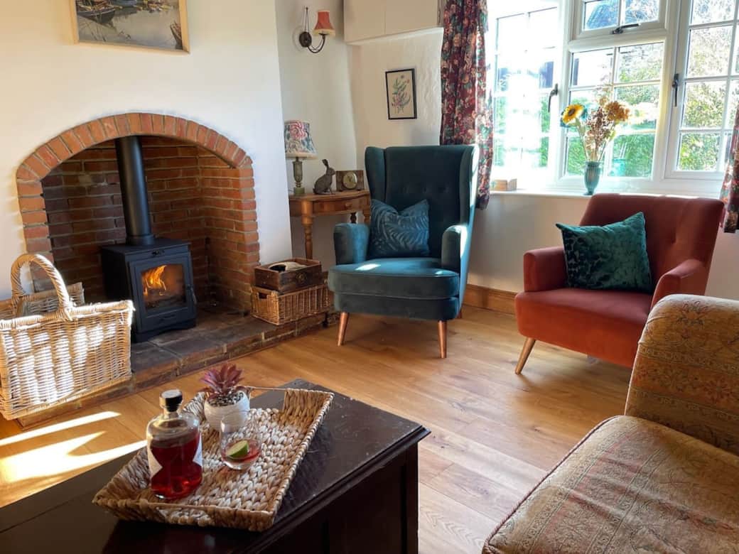 Living room with a lit log burner, exposed beams and a teal wing chair beside a sunlit window